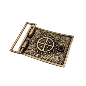 May include: A rectangular belt buckle made of antiqued brass-toned metal. The surface features a textured, cracked pattern with a central circular motif containing a cross and small dots. It includes a hinged bar and a small stud for fastening.