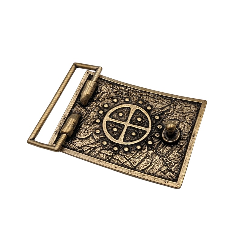 May include: A rectangular belt buckle made of antiqued brass-toned metal. The surface features a textured, cracked pattern with a central circular motif containing a cross and small dots. It includes a hinged bar and a small stud for fastening.