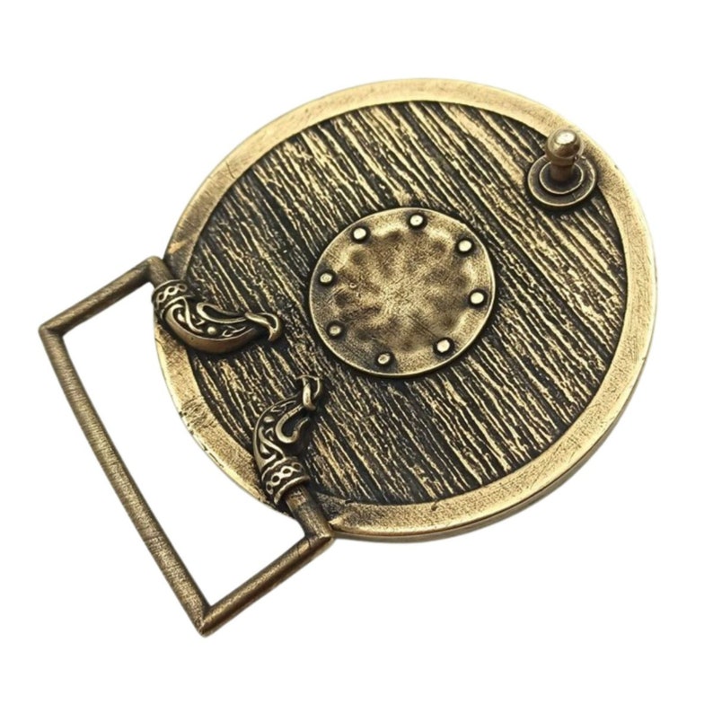 May include: A gold-toned metal belt buckle featuring a round shield design. The shield has a textured wood grain pattern and a smaller circular centre with rivets. The buckle has a metal loop for attaching a belt.