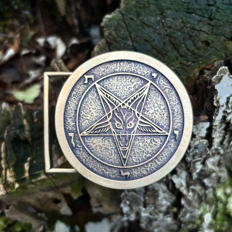 May include: A circular brass belt buckle featuring a raised pentagram design with a central goat head motif. The metal surface has a textured, antiqued finish with Hebrew characters engraved along the inner circular border of the emblem.