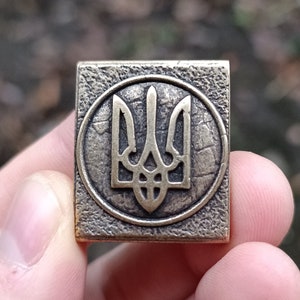 May include: A square-shaped metal pin with a textured surface. The pin features a gold-colored Ukrainian trident symbol inside a circle.