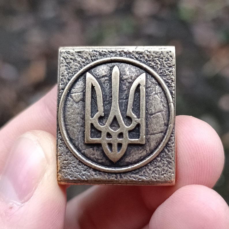 May include: A square-shaped metal pin with a textured surface. The pin features a gold-colored Ukrainian trident symbol inside a circle.