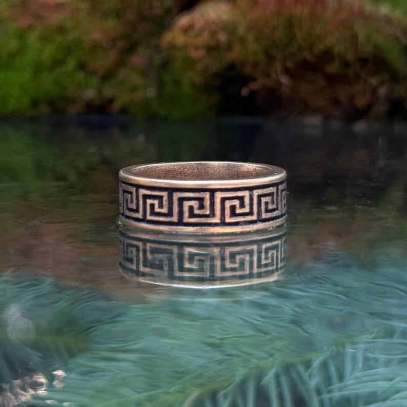 May include: A metallic band ring featuring a continuous black Greek key pattern engraved around the exterior surface. The ring has a smooth, polished finish and is captured with a clear reflection on the surface beneath it, showing the design.