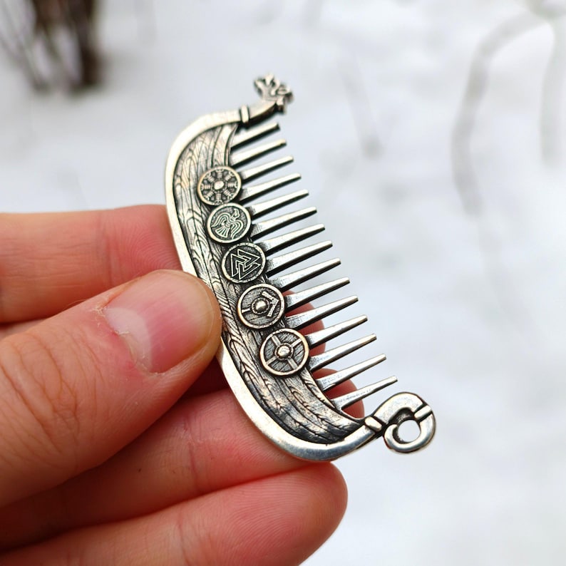 A silver plated comb with a Viking ship Drakkar design. The comb features a series of engraved shield emblems and a decorative flourish at the top. The comb's teeth are evenly spaced and extend from the ship's hull.