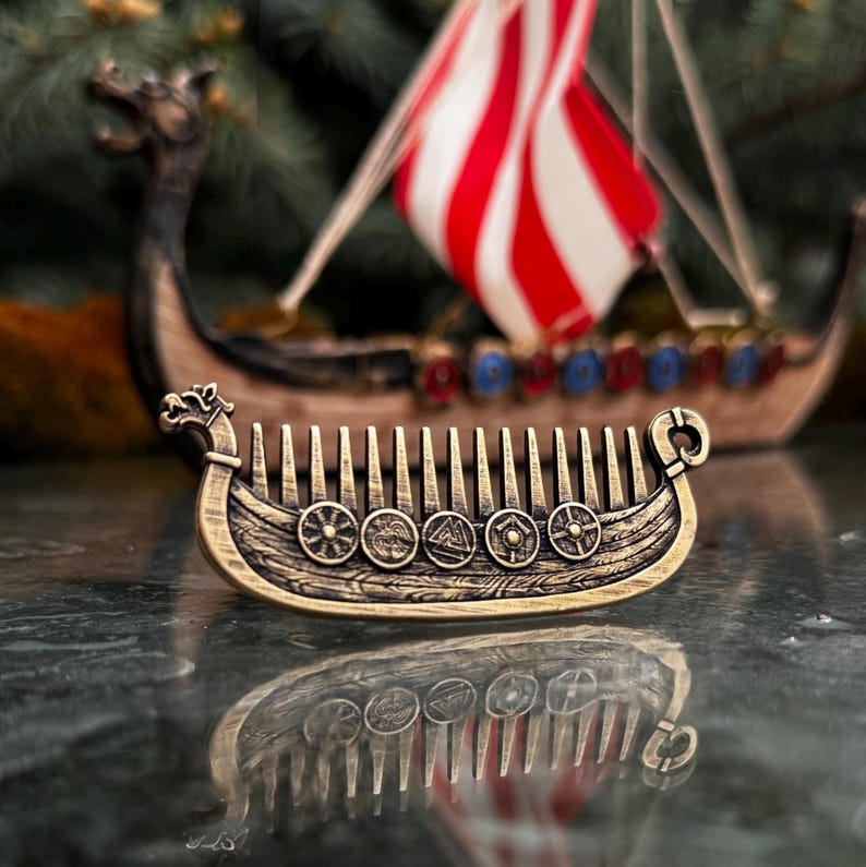A bronze norse beard comb shaped like a Drakkar, featuring intricate details and shield emblems. The comb's teeth are visible, and the design includes a dragon head at the front. A miniature Viking ship with a red and white striped sail is in the background.
