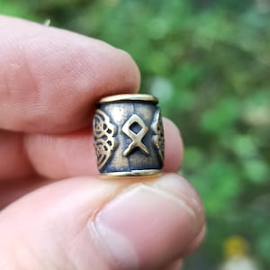 May include: A cylindrical metal bead with a dark, antiqued brass finish. The surface features a central engraved Othala rune symbol flanked by intricate, raised Celtic-style knotwork patterns on both sides, with smooth, polished rims.