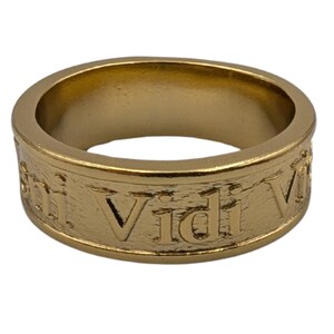 May include: A gold-toned metal ring with a textured surface featuring the embossed Latin phrase Veni Vidi Vici. The band has a consistent width and a smooth interior finish, showcasing raised lettering that wraps around the exterior circumference.