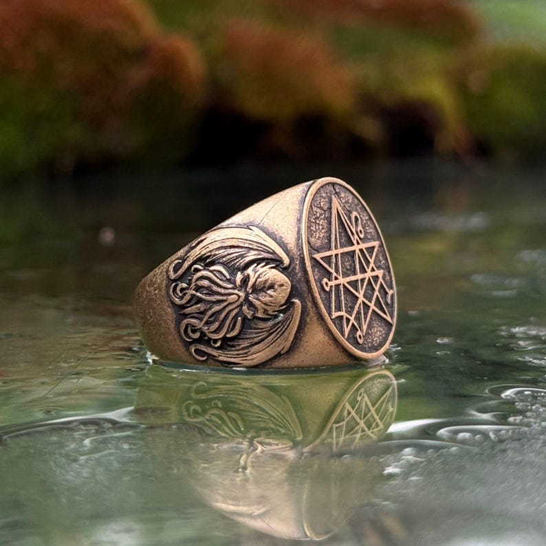 Może przedstawiać: Pierścień z metalu w kolorze brązu z okrągłą tarczą, na kt&oacute;rej wygrawerowano geometryczny symbol gwiazdy. Bok pierścienia zdobi szczeg&oacute;łowy relief przedstawiający stworzenie z mackami i skrzydłami, wykończony postarzaną, antyczną patyną.