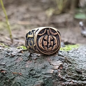 Puede incluir: Un anillo de tono dorado con un dise&ntilde;o detallado que presenta una calabaza tallada con una cara espeluznante. El anillo tiene el n&uacute;mero "13" en el lateral.