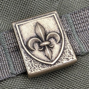 May include: A bronze-colored metal buckle with a fleur-de-lis design. The buckle is square-shaped and has a textured surface.