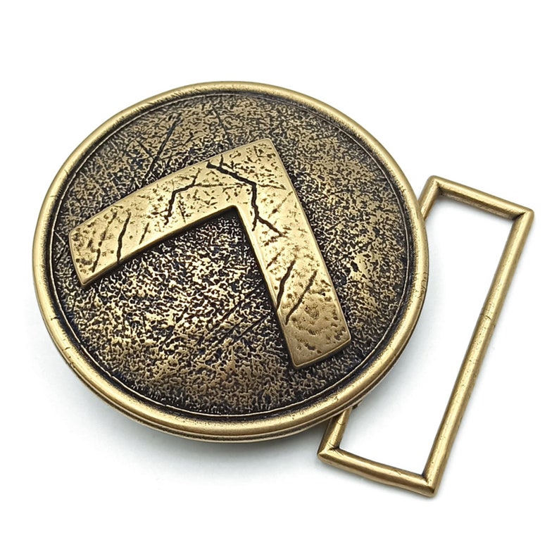 May include: A circular brass belt buckle with an antiqued, textured finish. The centre features a raised, V-shaped Spartan lambda symbol with a distressed, cracked surface. A rectangular loop is attached to the right side of the round buckle.