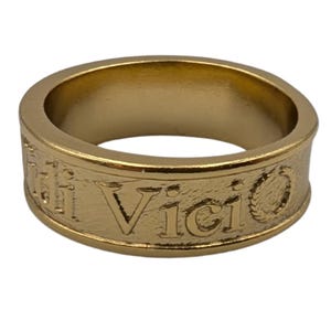 May include: A gold-toned metal band ring featuring a textured surface with the embossed text Veni Vidi Vici. The ring has a smooth, polished interior and a slightly raised rim along the top and bottom edges, creating a classic, uniform circular shape.