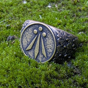 May include: A dark metallic ring featuring a circular face with an engraved Awen symbol consisting of three vertical rays topped by three dots. The band is decorated with a detailed relief pattern of oak leaves and acorns across the outer surface.
