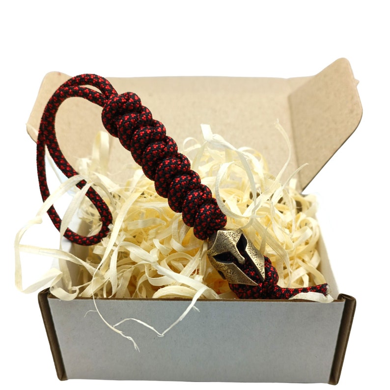 May include: A red and black paracord bracelet with a brass Spartan helmet charm. The bracelet is in a small cardboard box with wood shavings.