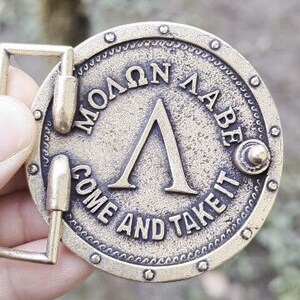 May include: A circular brass-toned metal belt buckle featuring a raised Greek Lambda symbol in the centre. The text "&Mu;&Omicron;&Lambda;&Omega;&Nu; &Lambda;&Alpha;&Beta;&Epsilon;" is embossed above the symbol, and the text "COME AND TAKE IT" is embossed below, surrounded by a studded border.