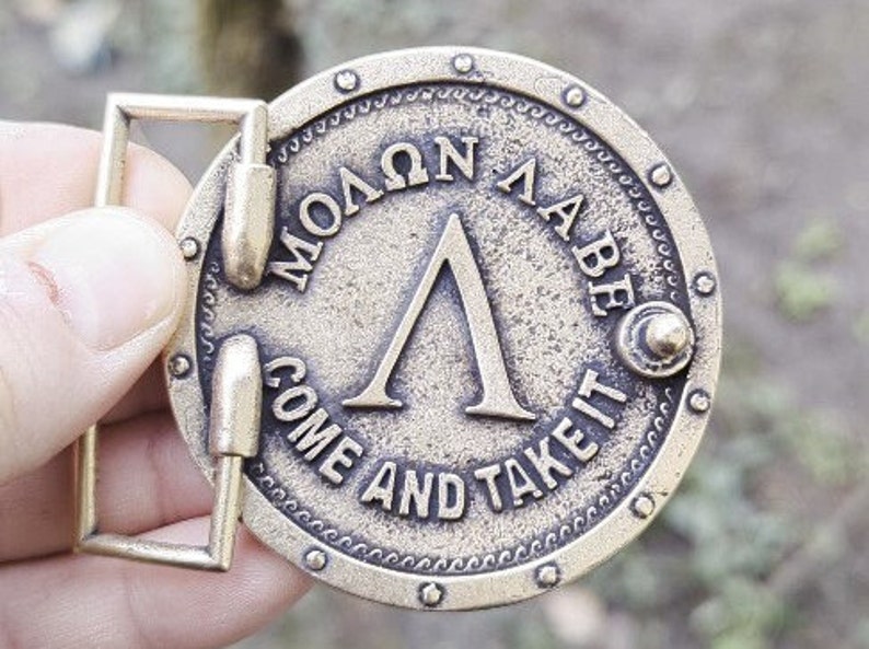 May include: A circular brass-toned metal belt buckle featuring a raised Greek Lambda symbol in the centre. The text "&Mu;&Omicron;&Lambda;&Omega;&Nu; &Lambda;&Alpha;&Beta;&Epsilon;" is embossed above the symbol, and the text "COME AND TAKE IT" is embossed below, surrounded by a studded border.
