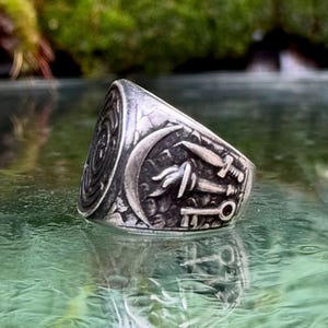 May include: A silver-toned metal ring featuring an oxidized finish with intricate embossed details. The design includes a crescent moon, a flaming torch, a dagger, and a key, all set against a textured background with a circular side emblem.