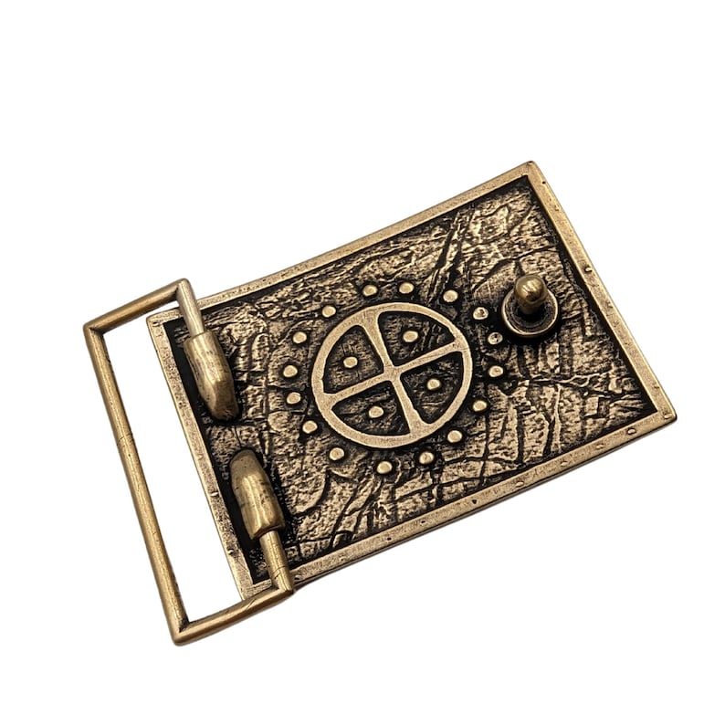 May include: A rectangular brass-toned belt buckle featuring a textured, weathered surface. The center displays a circular emblem with a cross and four dots, surrounded by a ring of small raised dots. A metal loop and a small stud are attached.