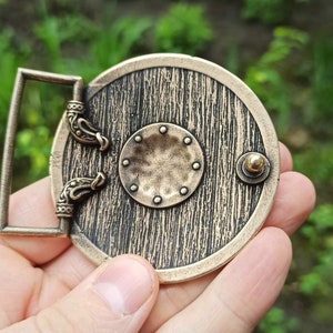 May include: A bronze belt buckle featuring a round shield design. The shield has a wood grain texture and a small round knob on the side. The buckle has two decorative loops on the side.