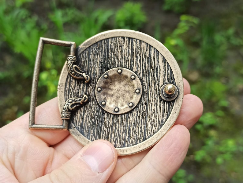 May include: A bronze belt buckle featuring a round shield design. The shield has a wood grain texture and a small round knob on the side. The buckle has two decorative loops on the side.