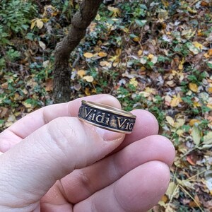 May include: A metallic band ring with a dark, weathered finish featuring the embossed Latin text Vidi Vici in a serif font. The ring has a flat profile and a textured surface, showcasing a contrast between the raised letters and the darkened background.