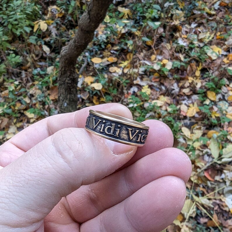 May include: A metallic band ring with a dark, weathered finish featuring the embossed Latin text Vidi Vici in a serif font. The ring has a flat profile and a textured surface, showcasing a contrast between the raised letters and the darkened background.