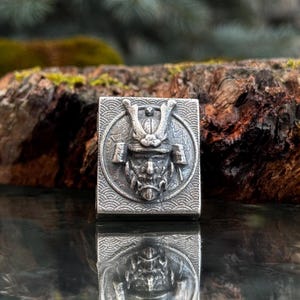 Könnte beinhalten: Eine rechteckige, silberfarbene Metallperle mit einem erhabenen Relief eines Samurai-Helms und einer Maske, zentriert in einem kreisförmigen Rahmen. Der Hintergrund der quadratischen Fläche ist mit einem Wellenmuster verziert.