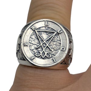 May include: A silver-toned metal ring featuring a circular face with a textured background. The center displays a sigil of Lucifer, surrounded by the letters L, U, C, I, F, E, R engraved along the circular border. The band has a rugged, weathered finish.