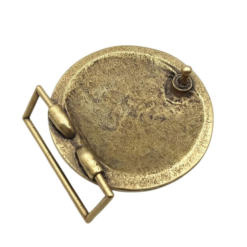 May include: A circular brass-toned belt buckle with a textured, weathered finish. The back of the buckle features a rectangular loop for the belt strap and a small, upright metal prong designed to secure the belt through a hole.