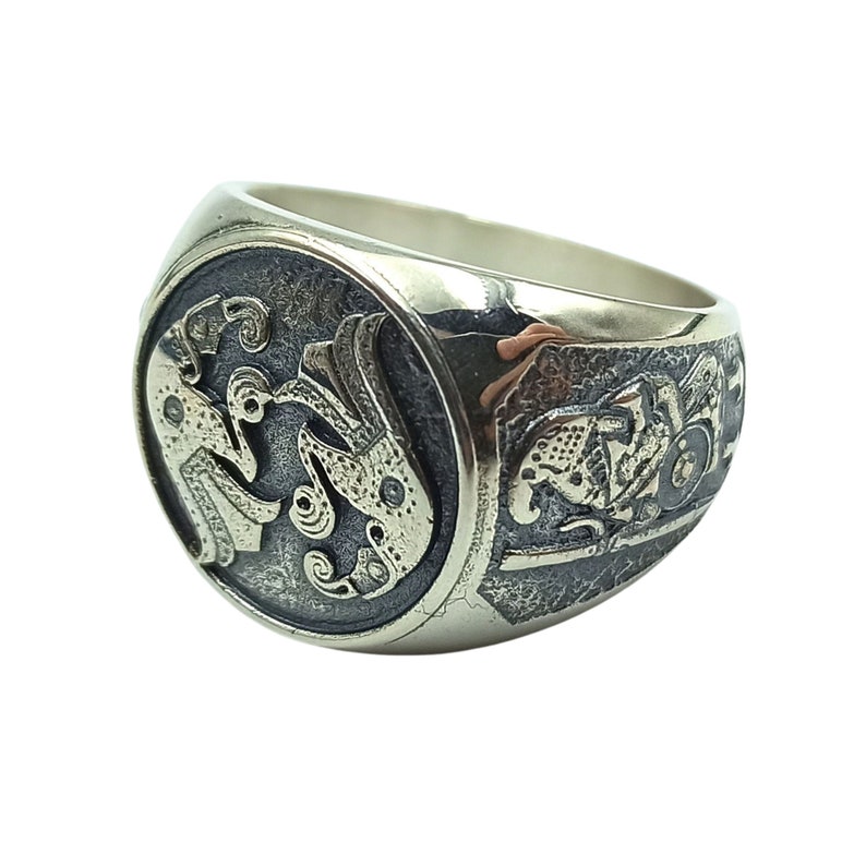 May include: A silver ring with an intricate design featuring two stylized figures and a detailed background. The ring has a wide band and a rounded top.
