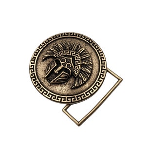 May include: A circular bronze-toned belt buckle featuring a raised relief of a Spartan helmet in the centre. The outer edge of the circular design is decorated with a repeating Greek key pattern. A rectangular metal loop is attached to the side.