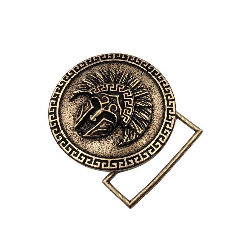 May include: A circular bronze-toned belt buckle featuring a raised relief of a Spartan helmet in the centre. The outer edge of the circular design is decorated with a repeating Greek key pattern. A rectangular metal loop is attached to the side.