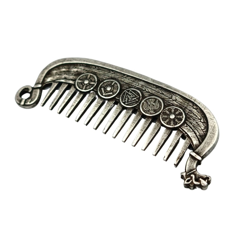 A silver-colored metal beard comb shaped like a Viking longship Drakkar. The comb features intricate detailing, including shields with various symbols. The comb's design includes a loop at one end and a decorative element resembling a ship's prow at the other.