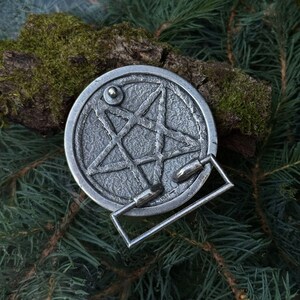 May include: A circular silver-toned metal belt buckle featuring a raised five-pointed star design within a textured border. The buckle includes a rectangular frame attachment and a small circular stud on the upper left side of the star motif.