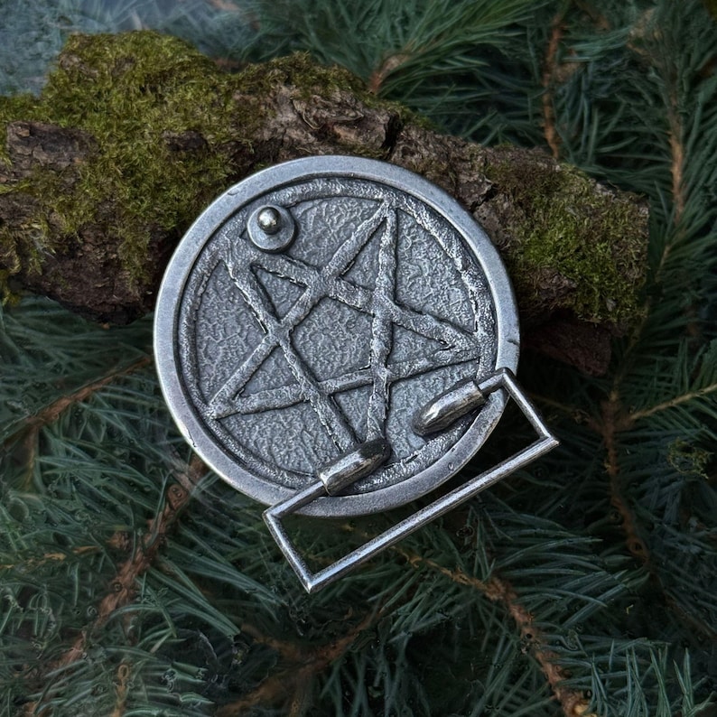 May include: A circular silver-toned metal belt buckle featuring a raised five-pointed star design within a textured border. The buckle includes a rectangular frame attachment and a small circular stud on the upper left side of the star motif.