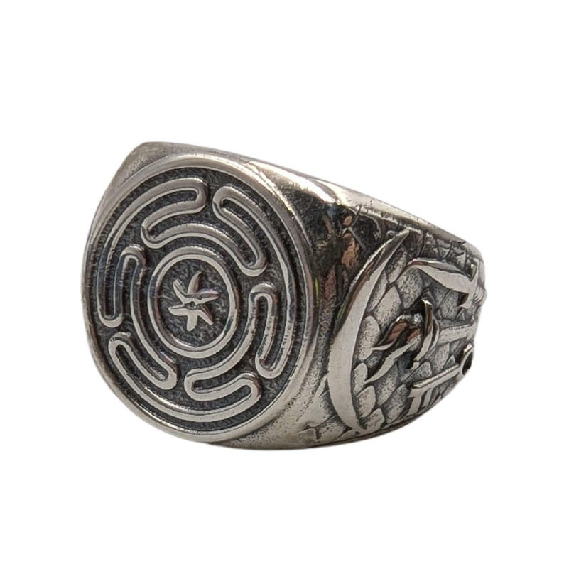May include: A silver-toned metal ring featuring a circular face with an embossed labyrinthine pattern and a central star-like symbol. The sides of the band display intricate relief carvings of a crescent moon and a sword against a textured background.