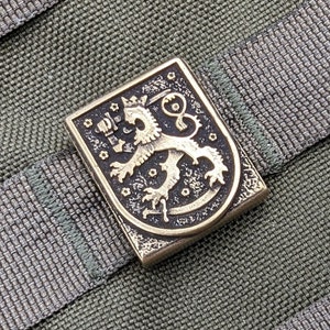 Op de afbeelding: Een gouden metalen pin met het wapen van Finland. Het wapen toont een leeuw met een kroon op zijn kop en een zwaard in zijn poot. De leeuw staat op een schild met een zwarte achtergrond en gouden sterren.