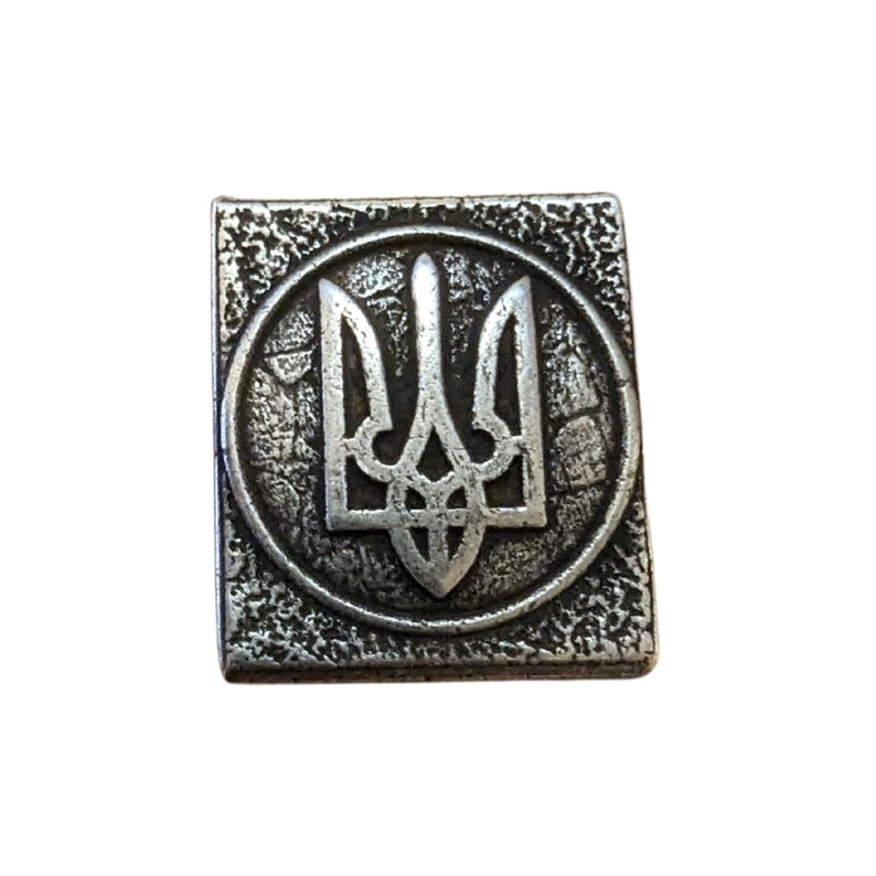 May include: A silver metal pin with a textured surface and a raised design of the Ukrainian trident, a three-pronged spear.