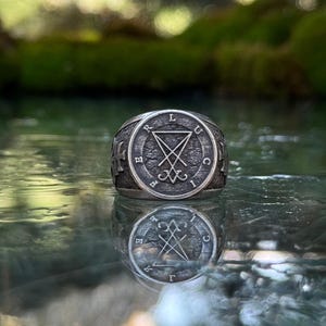May include: A silver-toned metal ring featuring a circular face with a textured background. The center displays a sigil of Lucifer, surrounded by the letters L, U, C, I, F, E, R arranged in a circle. The sides of the ring band feature a cross emblem.