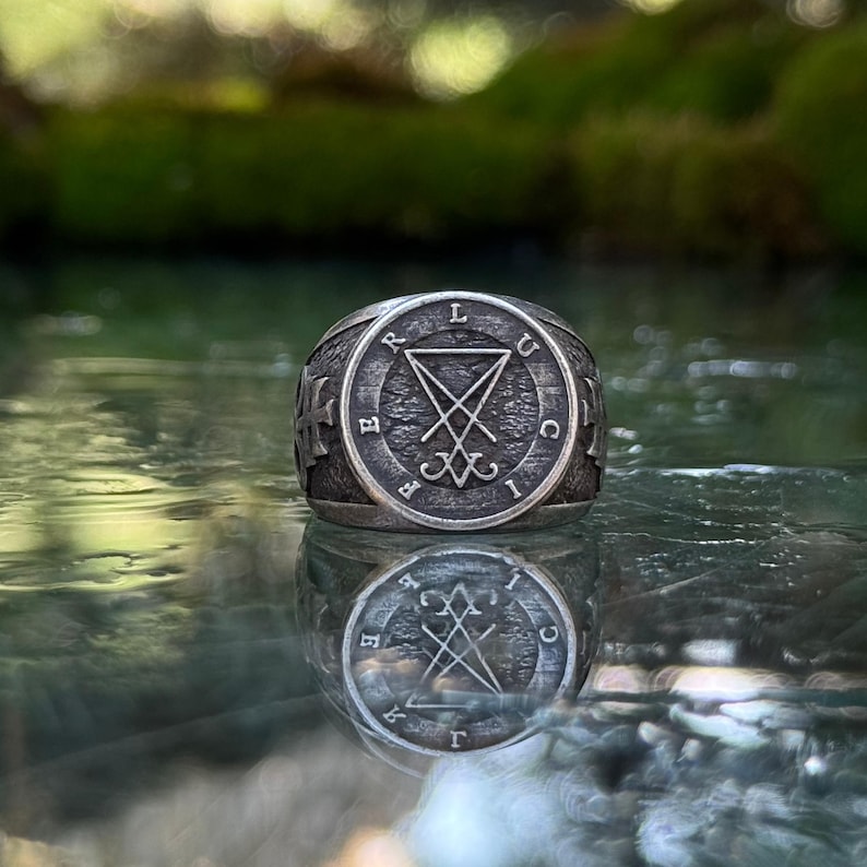 May include: A silver-toned metal ring featuring a circular face with a textured background. The center displays a sigil of Lucifer, surrounded by the letters L, U, C, I, F, E, R arranged in a circle. The sides of the ring band feature a cross emblem.