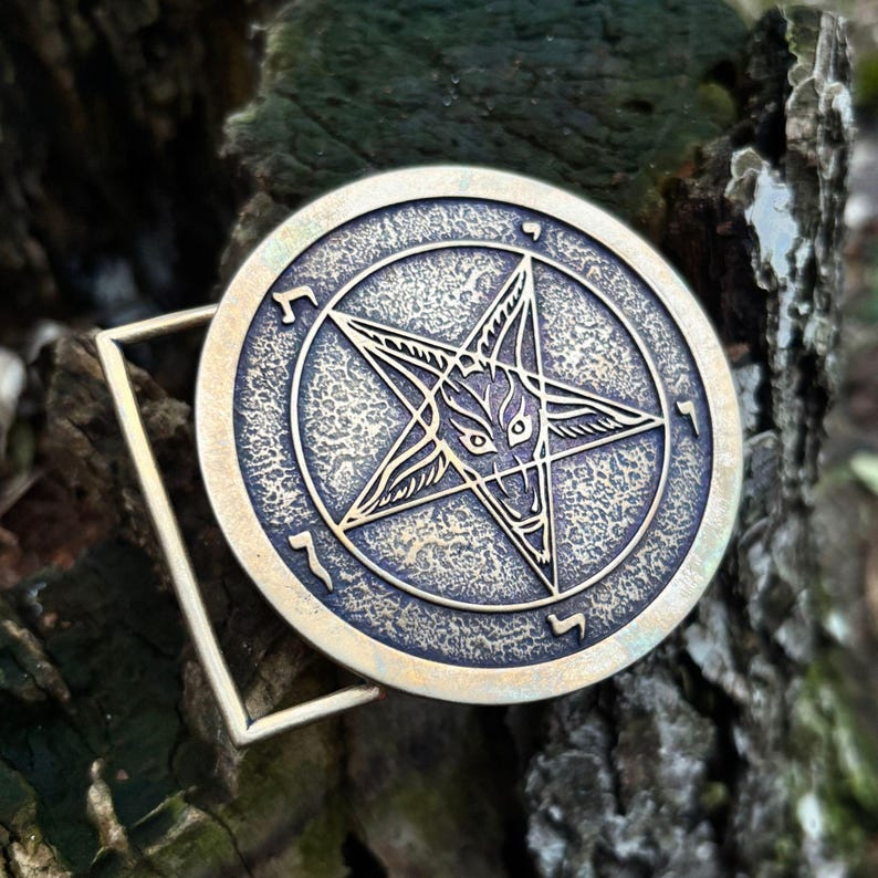 May include: A circular brass-toned metal belt buckle featuring a raised pentagram design with a central goat head motif. The surface has a textured, weathered finish with engraved symbols positioned between the points of the star within the circular border.