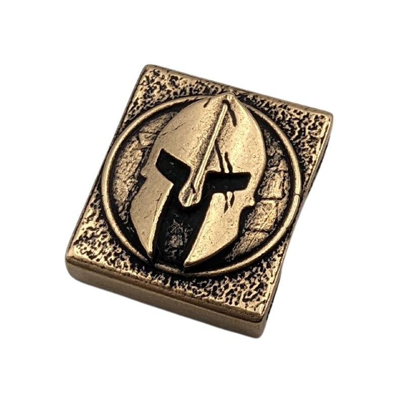 May include: A gold-colored metal square charm with a raised, detailed image of a Spartan helmet in black and gold.