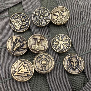 Pode incluir: Uma coleção de nove botões de metal com cor de latão antigo com vários símbolos vikings. Os desenhos incluem uma cabeça de carneiro, um escudo, um martelo e outros padrões intrincados. Os botões são redondos e têm uma aparência ligeiramente envelhecida.
