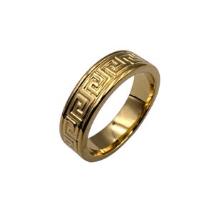 May include: A polished gold-tone ring featuring a continuous engraved Greek key pattern around the outer band. The metal surface has a reflective finish, highlighting the geometric interlocking lines that wrap around the entire circumference of the ring.