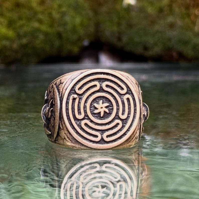 May include: A metallic ring featuring a circular face with an engraved labyrinth pattern and a central six-pointed star symbol. The band has a weathered, bronze-toned finish with intricate carvings on the sides, showing a reflective surface.