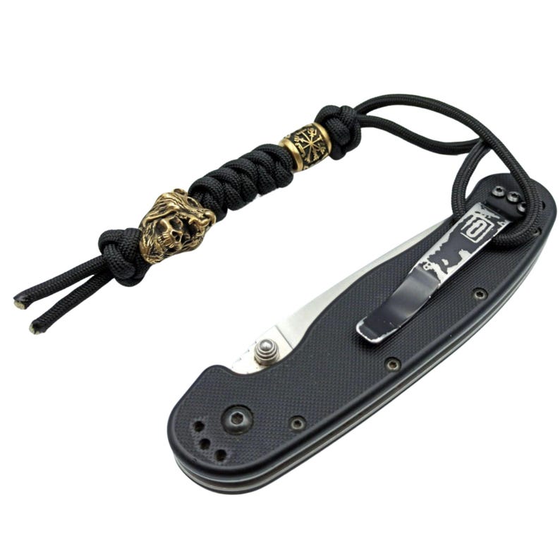 May include: A black folding knife with a black paracord lanyard featuring a skull bead and a metal bead with a symbol. The knife has a pocket clip.