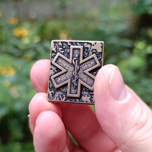 Pode incluir: Um alfinete de metal de cor bronze com um padr&atilde;o de camuflagem preto e branco. O alfinete apresenta um s&iacute;mbolo m&eacute;dico estilizado com uma cobra enrolada em torno de um cajado.