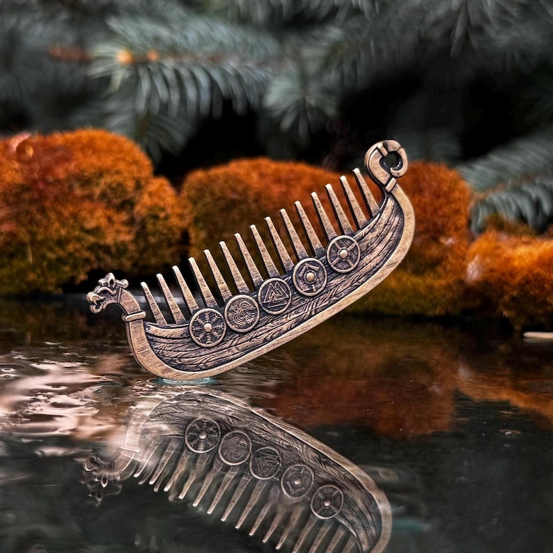 A bronze beard comb shaped like a Viking longship, featuring intricate details and circular shield designs. The comb is reflected in a dark, reflective surface, with a blurred background of greenery and moss.