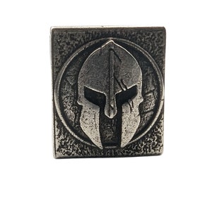 May include: A silver metal square with a raised image of a Spartan helmet in the center. The helmet is detailed with a crest and a face mask.