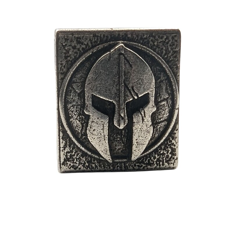 May include: A silver metal square with a raised image of a Spartan helmet in the center. The helmet is detailed with a crest and a face mask.